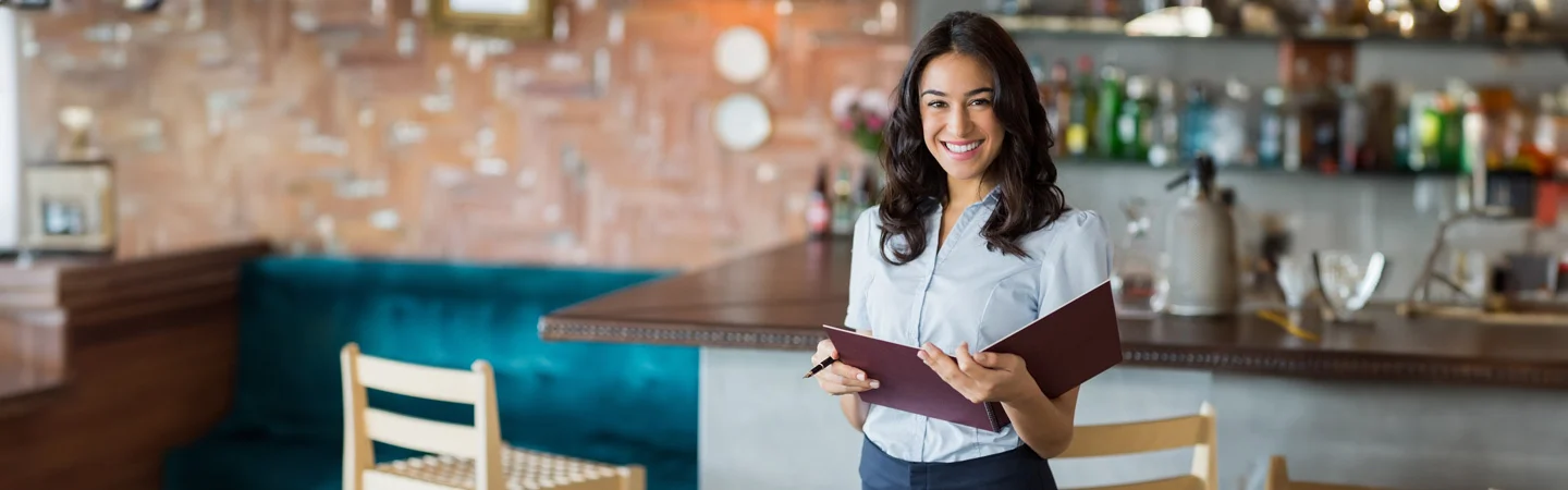 A smiling woman holding a menu stands in a stylish restaurant with a bar and wooden chairs in the background.