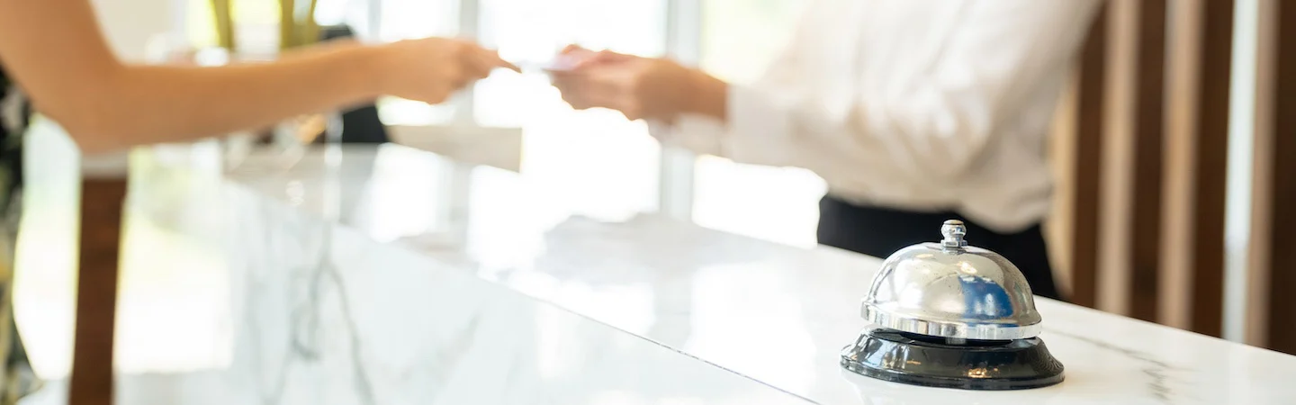 A person hands a card to a receptionist at a hotel front desk, with a service bell in the foreground.