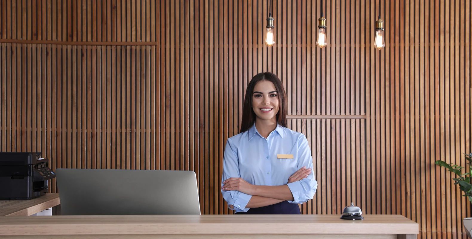 A receptionist in a blue shirt stands smiling with folded arms behind a desk, with wooden paneling and lights in the background.