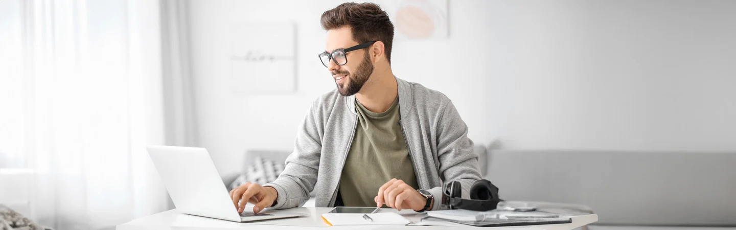 A man with glasses, wearing a gray cardigan, works on a laptop at a white desk with papers and a tablet.