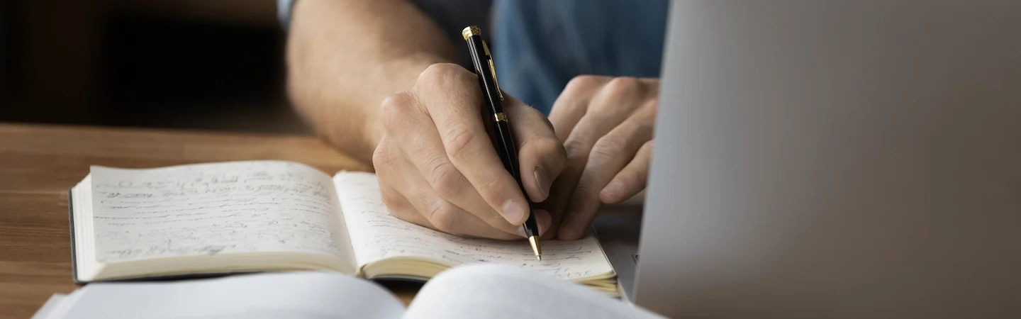Person writing notes in a notebook with a pen, laptop and an open book are on the desk.