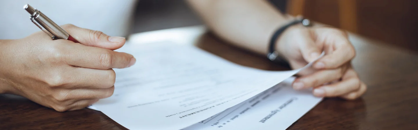 A person holding a pen and a document while reviewing or signing it, sitting at a table.
