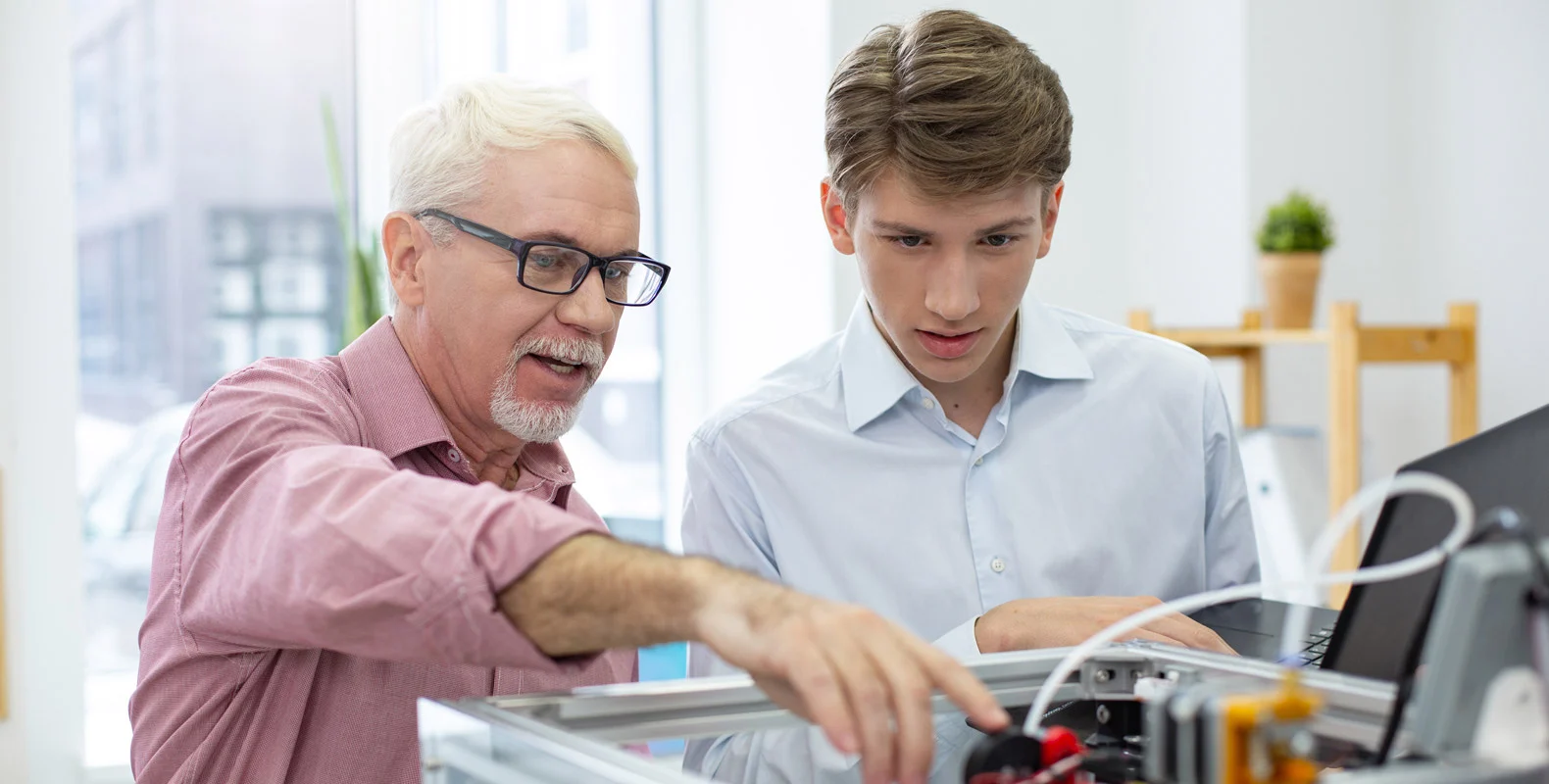 An older man and a younger man work together on a piece of machinery, both focused and discussing the task.
