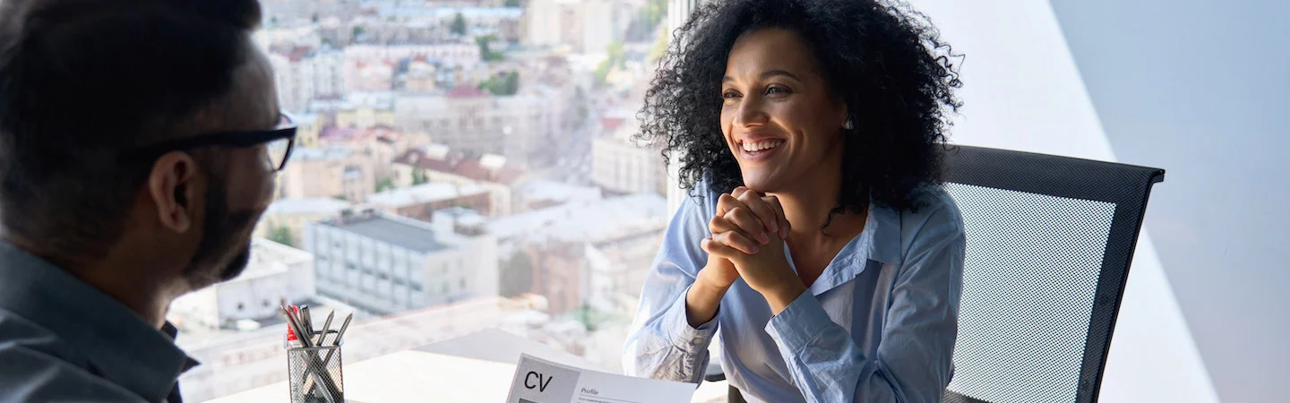 Woman smiling during an office interview, holding a resume. Bright cityscape visible through the large window behind them.