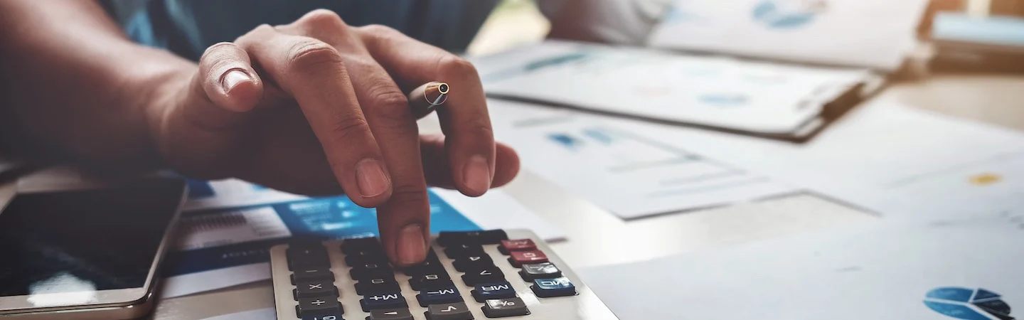 Close-up of a hand holding a pen and pressing a calculator with financial documents spread out on a desk.