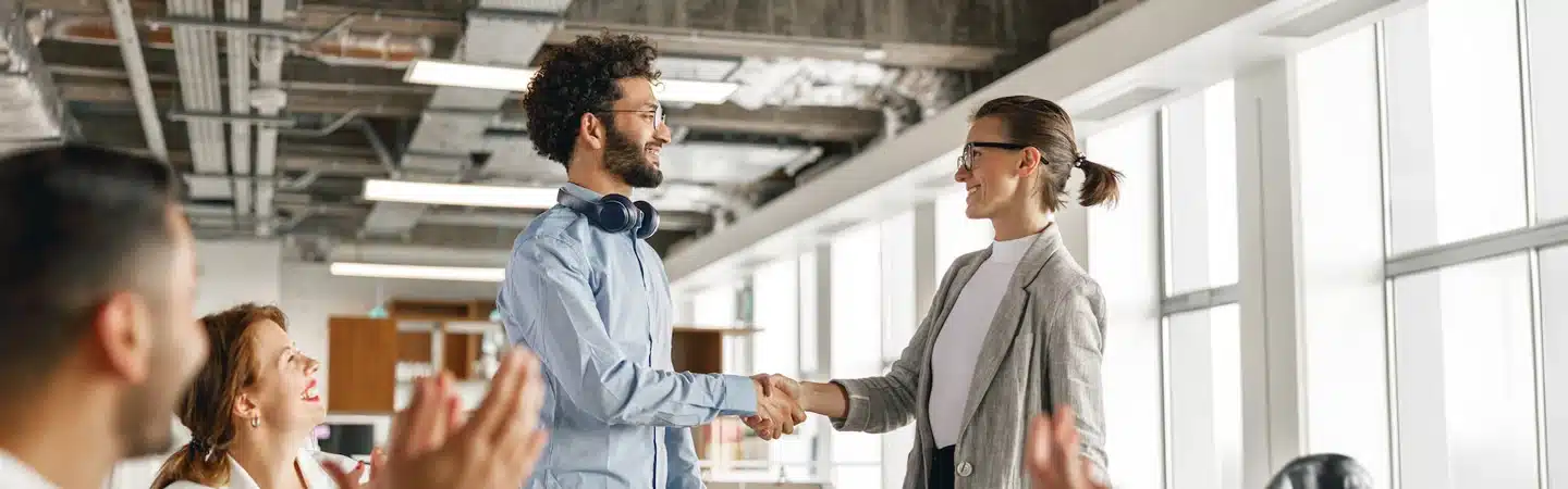 Two colleagues, a man and a woman, shaking hands in a modern office while others applaud in the background.