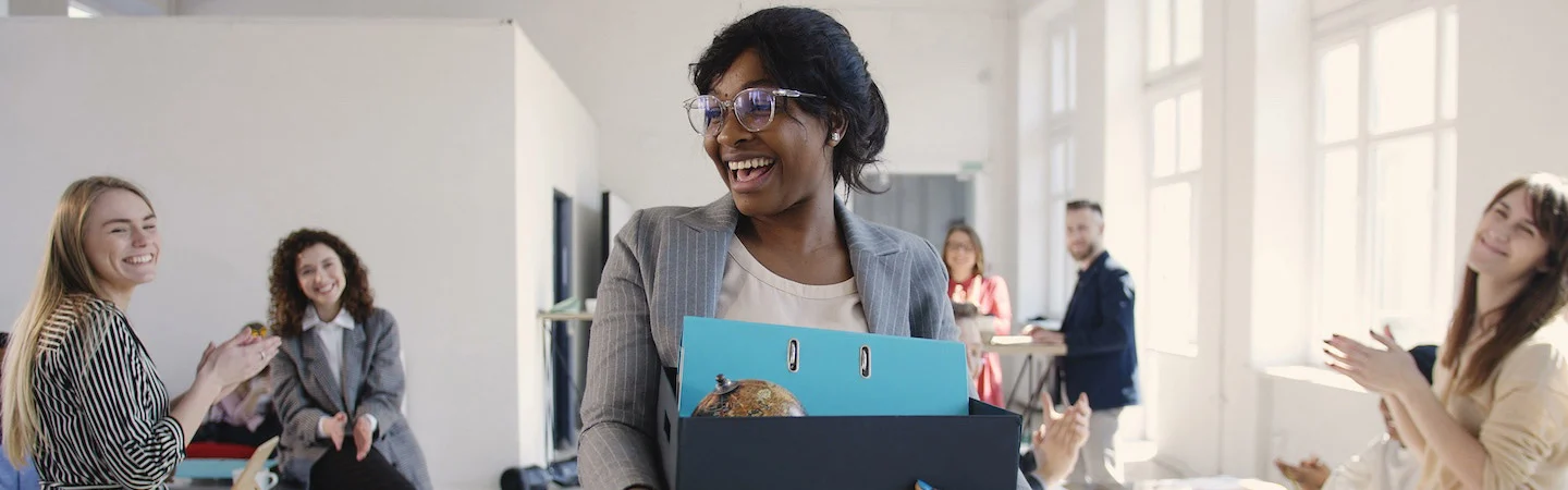 A smiling woman carrying a box is welcomed by colleagues in a bright office.