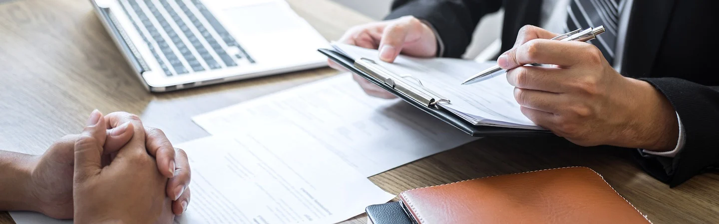 Two people in a business meeting; one holding a clipboard and pen, the other with clasped hands on the table.