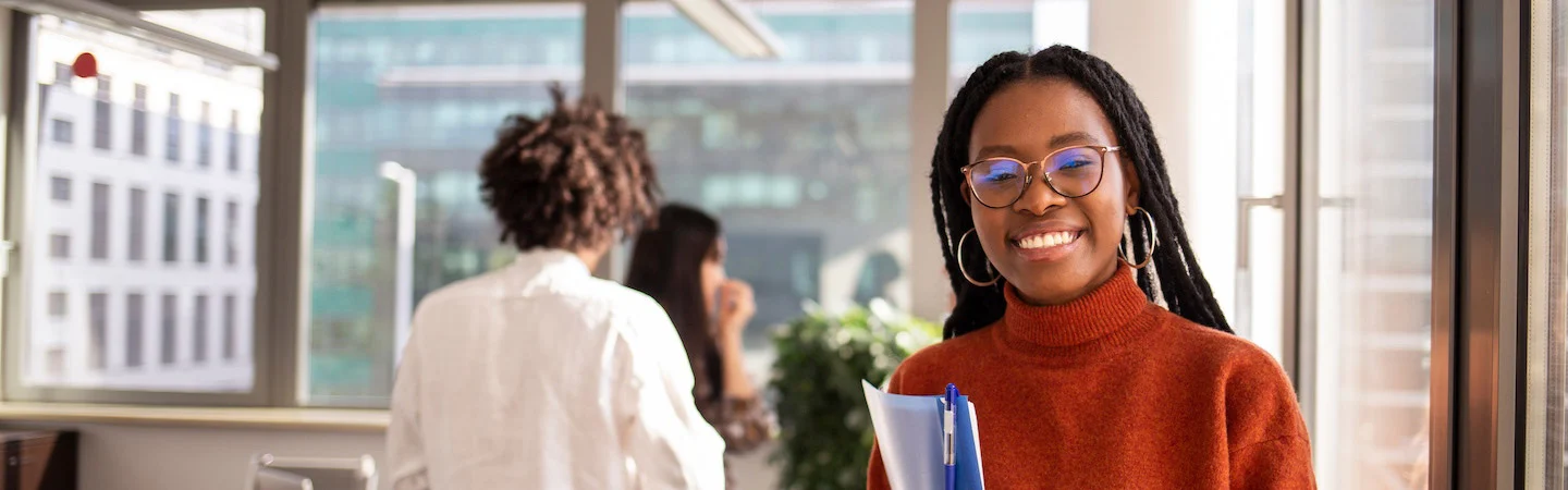 Smiling woman with glasses and braided hair holding files in a bright office; two people talking in the background.