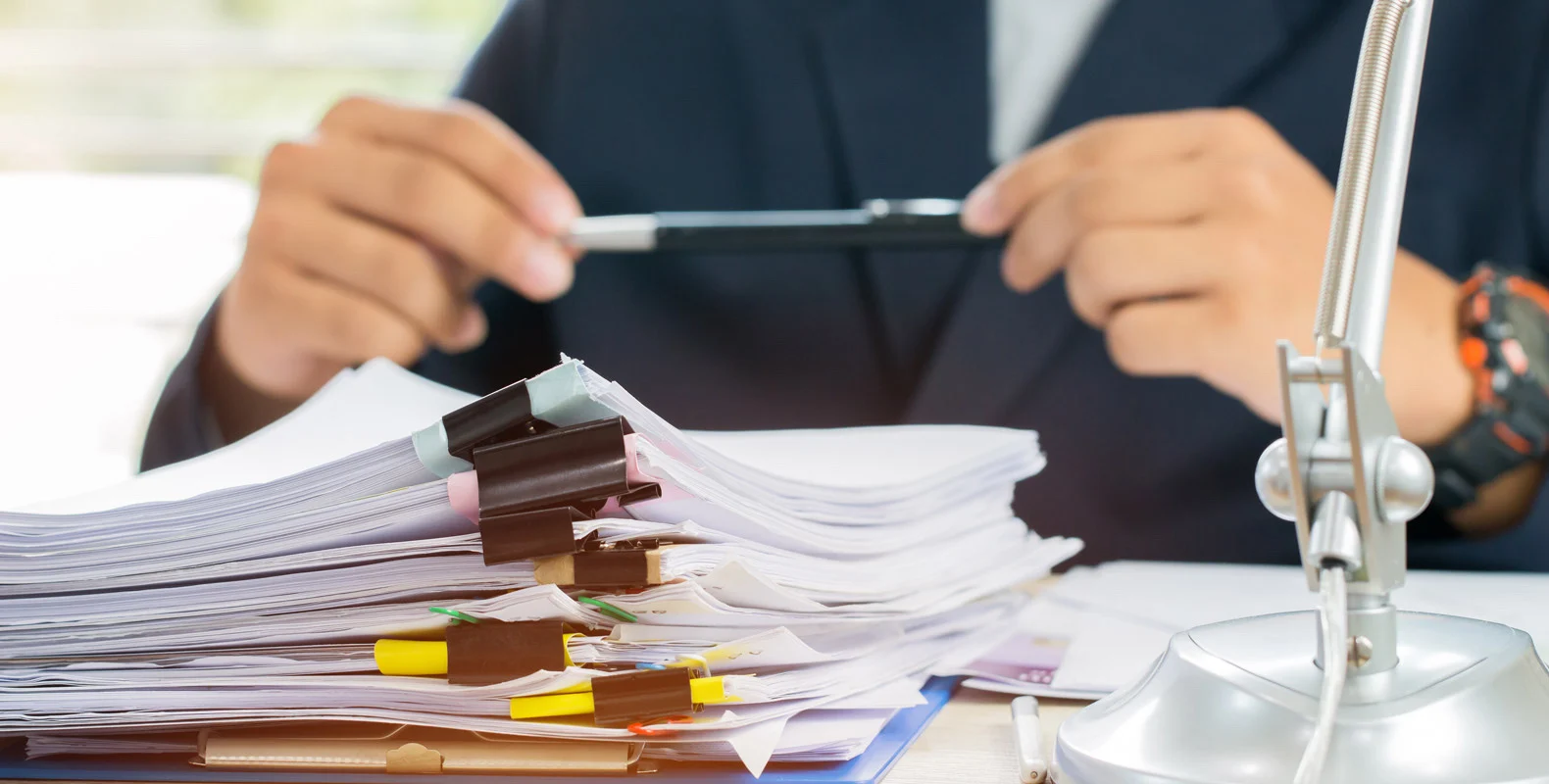 Person holding a pen, sitting at a desk with a large stack of documents clipped together and a desk lamp nearby.