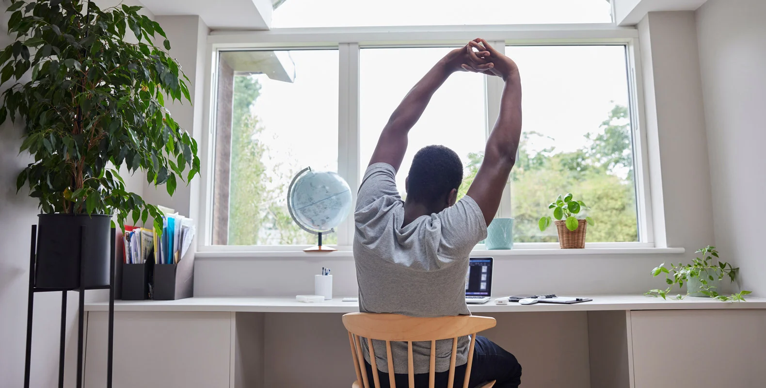Person stretching in front of a desk with a laptop, plants, and globe, facing a large window displaying greenery outside.