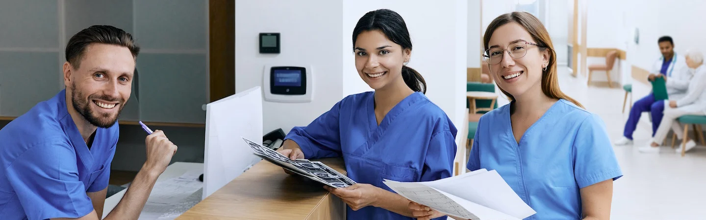 Three smiling medical professionals in scrubs hold clipboards at a hospital desk, with patients in the background.