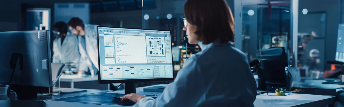 Person working on a computer, viewing complex data and coding in a modern lab setting with colleagues in the background.