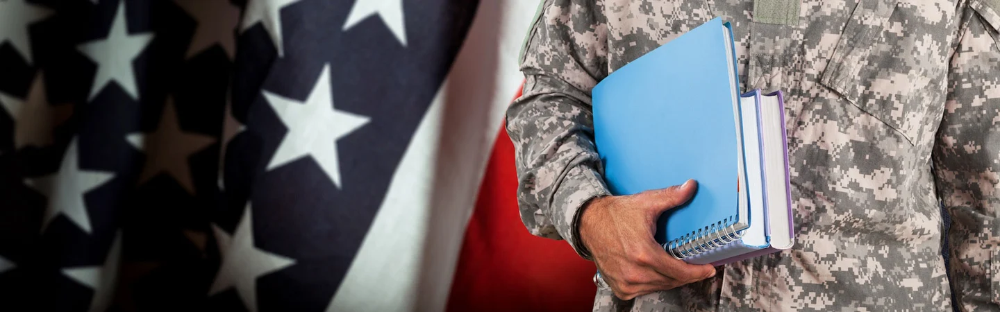 A person in military uniform holding notebooks, with a close-up of the American flag in the background.