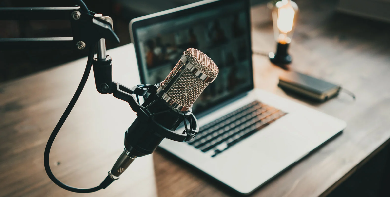 Close-up of a microphone on a stand in front of a laptop on a wooden desk, with a notebook and a light bulb in the background.