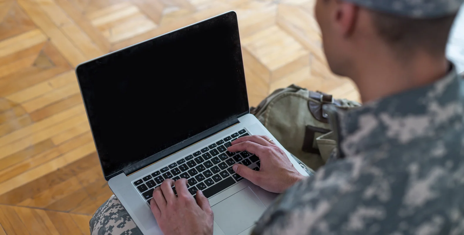 Person in military uniform typing on a laptop, sitting on a wooden floor with a backpack beside them.