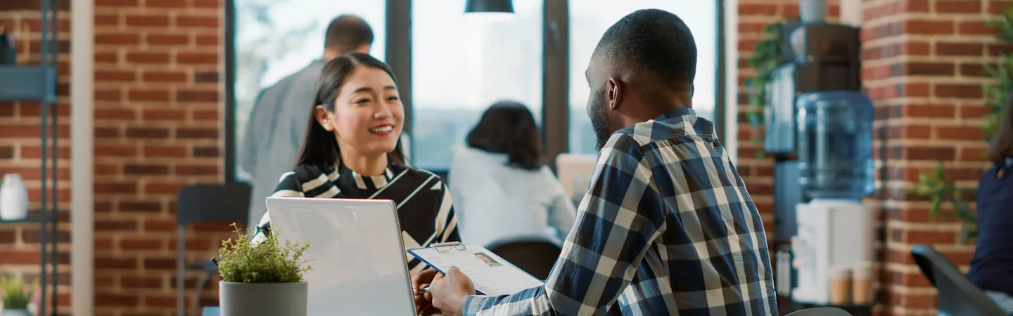Two people having a cheerful conversation at a desk in a busy office with brick walls and large windows.