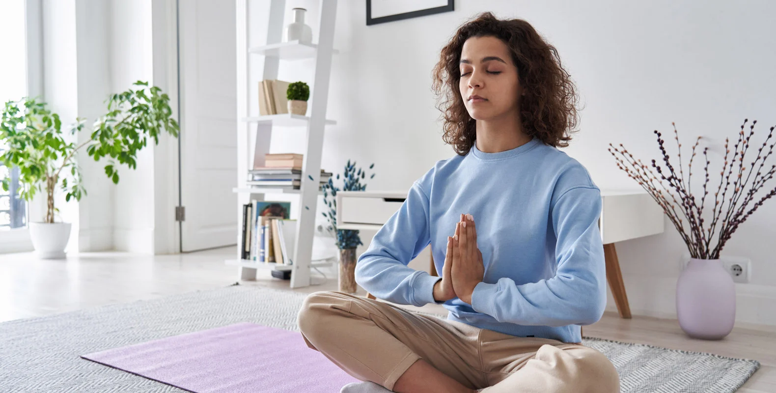 A person with curly hair is sitting cross-legged on a yoga mat in a peaceful room, meditating with eyes closed and hands together.