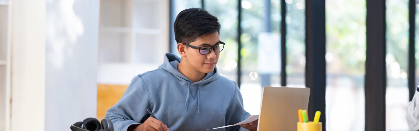 Person in a hoodie and glasses working at a laptop, holding papers, with a bright window in the background.