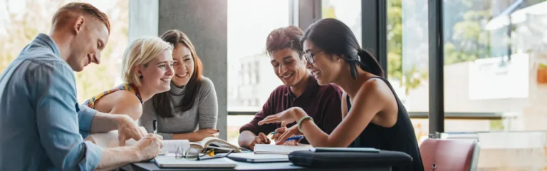 A group of five students sitting at a table, studying and smiling together in a well-lit room with large windows.