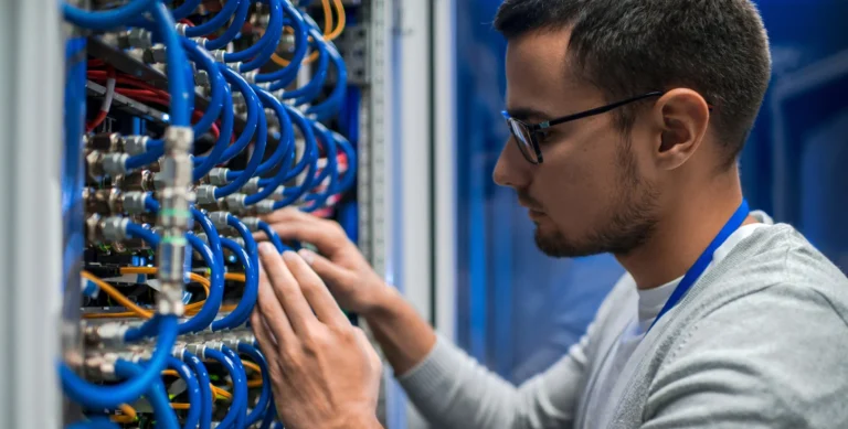 A man wearing glasses and a lanyard adjusts cables in a server room filled with blue and yellow wires.
