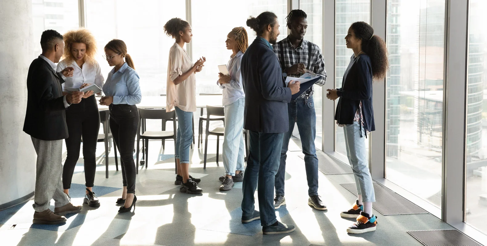 A group of people stand in pairs, chatting and holding documents in a modern office space with large windows.