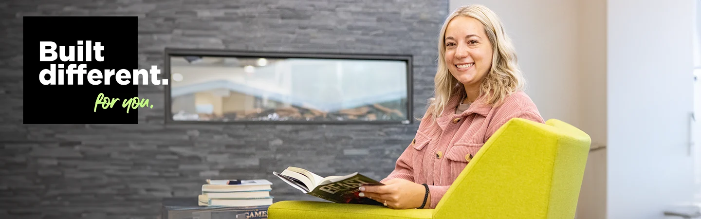 A woman sits on a green chair reading a book, smiling. Text reads "Built different. for you." Background features a modern room.