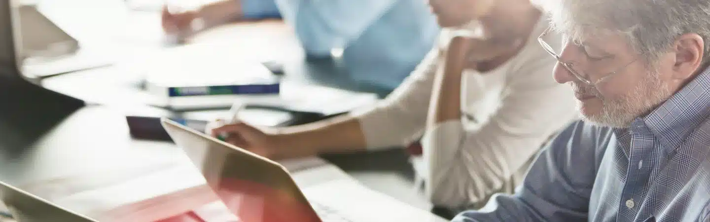 People working on laptops and taking notes at a table, focusing on a task in a bright, modern workspace.