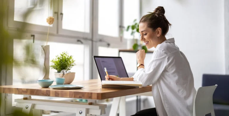 A woman sits at a wooden desk near a window, smiling and writing in a notebook with a laptop open in front of her.