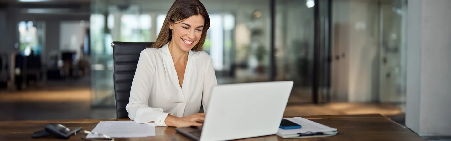 Woman in a white blouse working on a laptop at a desk in a modern office, smiling and appearing focused.