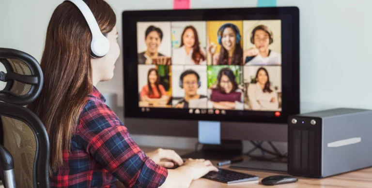 Woman wearing headphones participating in a video conference on a computer with eight people on the screen.