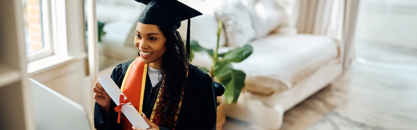 A graduate in cap and gown holds a rolled diploma, smiling in a brightly lit room with a sofa and potted plant.