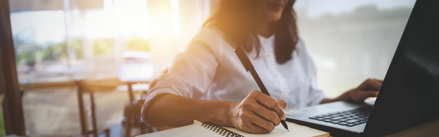 A person writes in a notebook while using a laptop, with sunlight streaming through a window in the background.