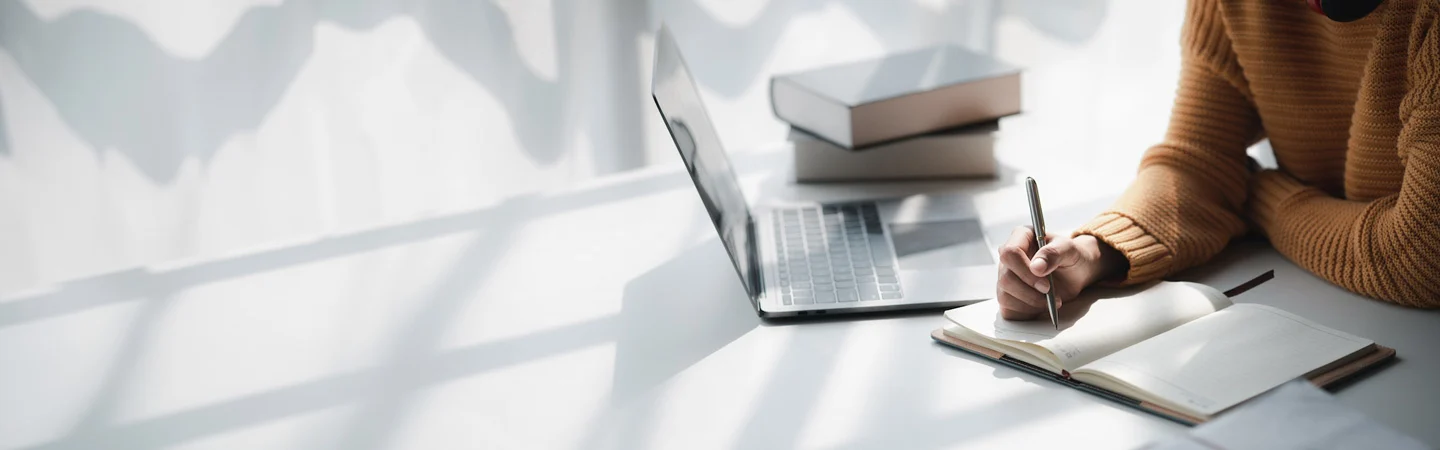 Person in a yellow sweater writing in a notebook beside a laptop and a stack of books on a sunlit desk.