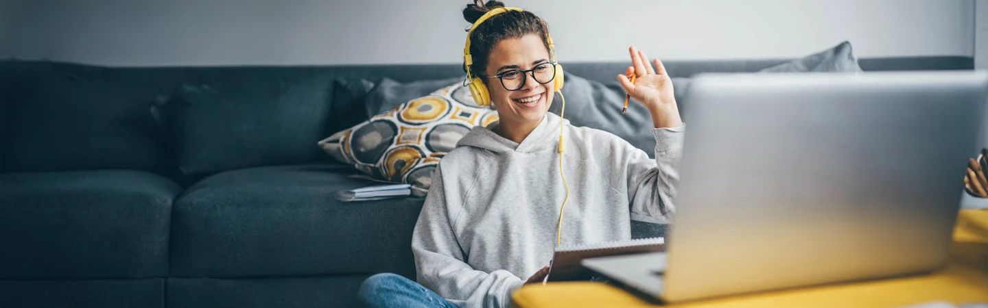 Person wearing headphones and glasses, smiling and waving at a laptop in a comfortable, modern living room.