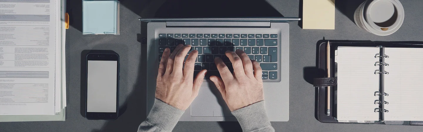 Hands typing on a laptop keyboard on a desk with documents, a smartphone, coffee, and an open planner.