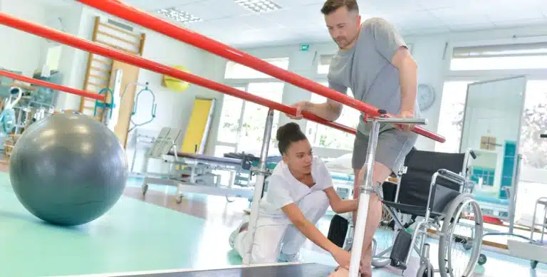 Occupational Therapy Assistant helps a man walk with parallel bars in a rehabilitation center, wheelchair and exercise equipment nearby.