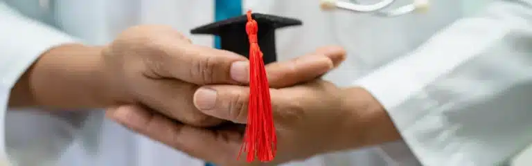 Person in a white coat holding a small graduation cap with a red tassel in their hands.