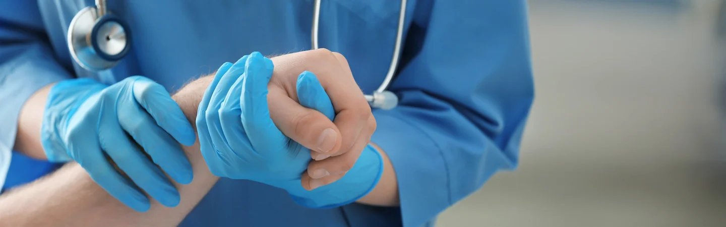 Close-up of a healthcare professional in blue scrubs and gloves checking the pulse of a patient's wrist.