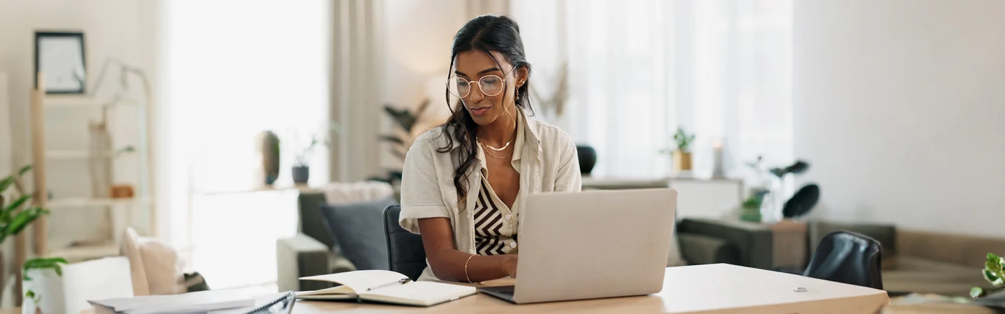 A woman sits at a desk with a laptop, reading a notebook. She wears glasses and is in a bright, well-decorated room.