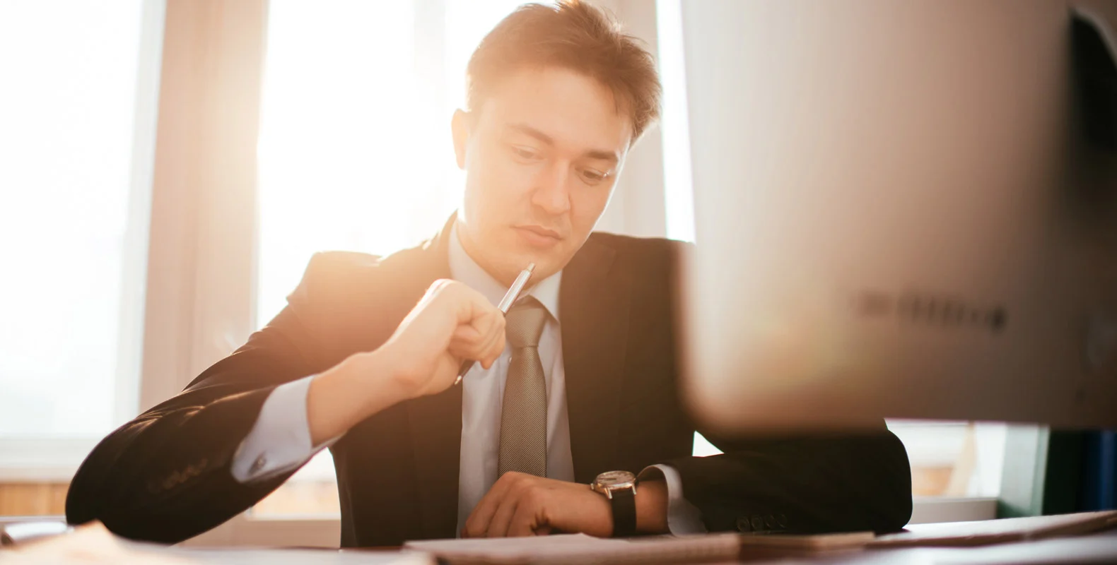 Man in a suit working with a computer, thoughtfully holding a pen in his hand with sunlight streaming in behind him.