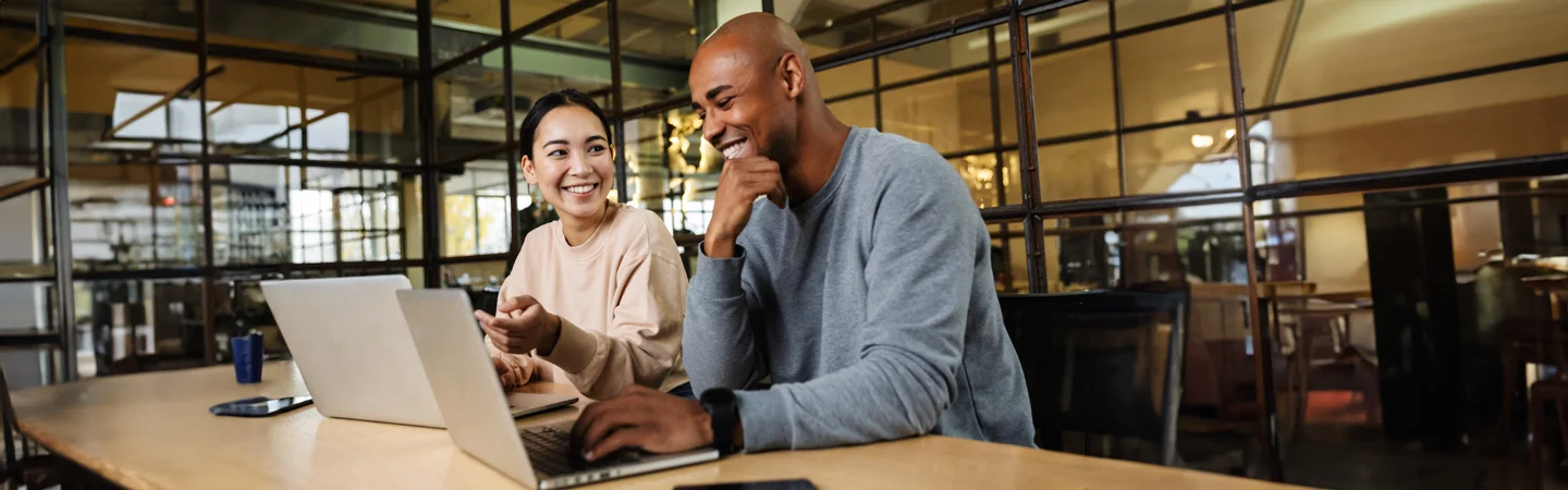 Two people smiling and working on laptops in a modern office with glass walls and shelves in the background.
