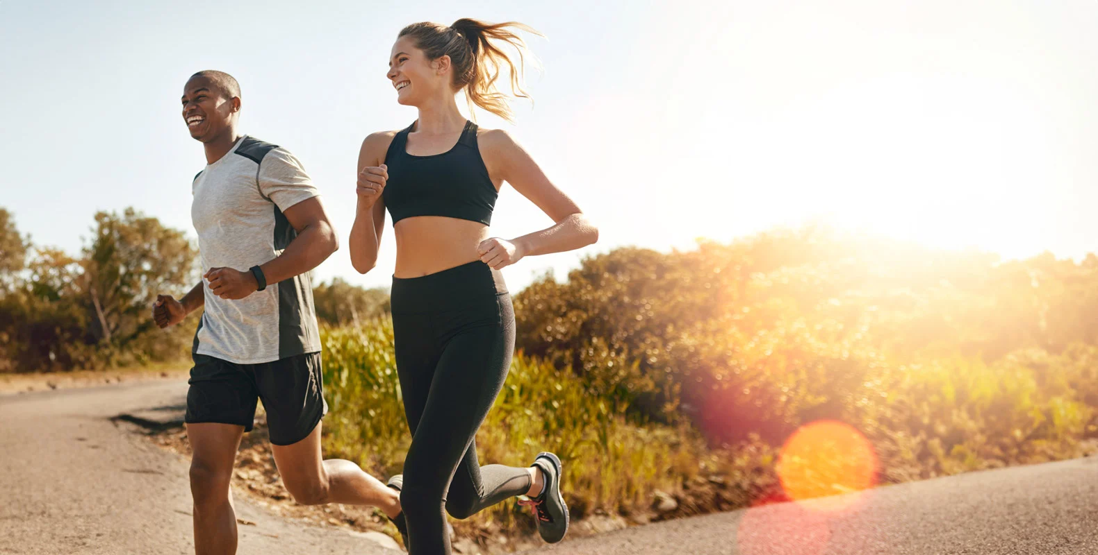 A man and a woman jogging together on a sunny day, with trees and bushes in the background.