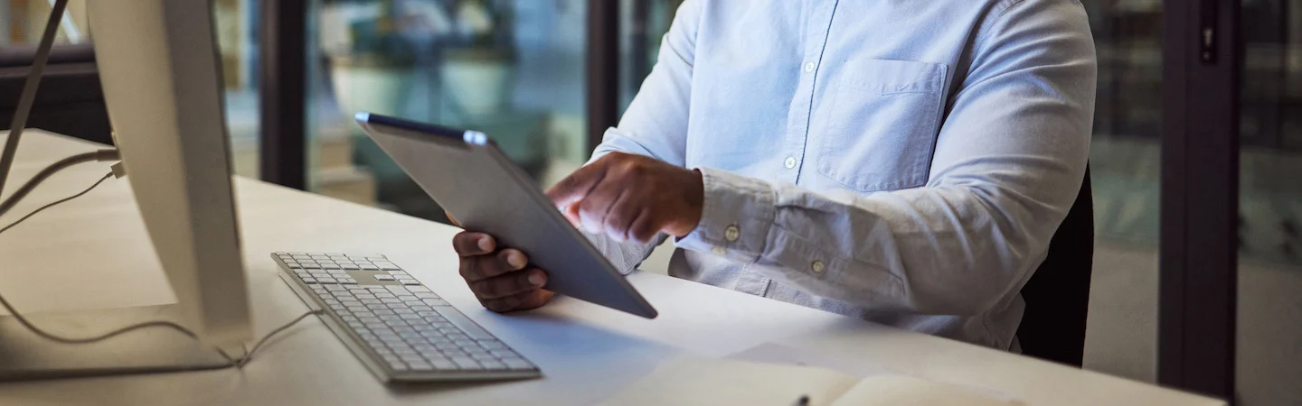 Person in a light blue shirt using a tablet at a desk with a keyboard and a computer monitor in a modern office setting.