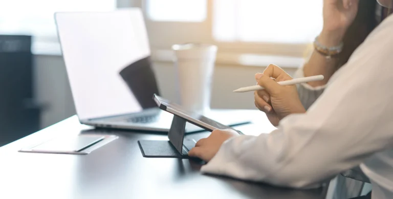 Person using a tablet with a stylus at a desk, with a laptop and a cup in the background.