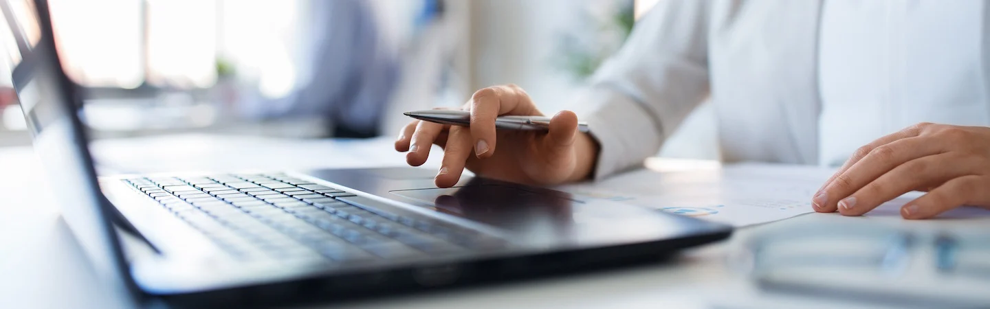 Close-up of a person's hand holding a pen and using a laptop touchpad, with documents on the desk.