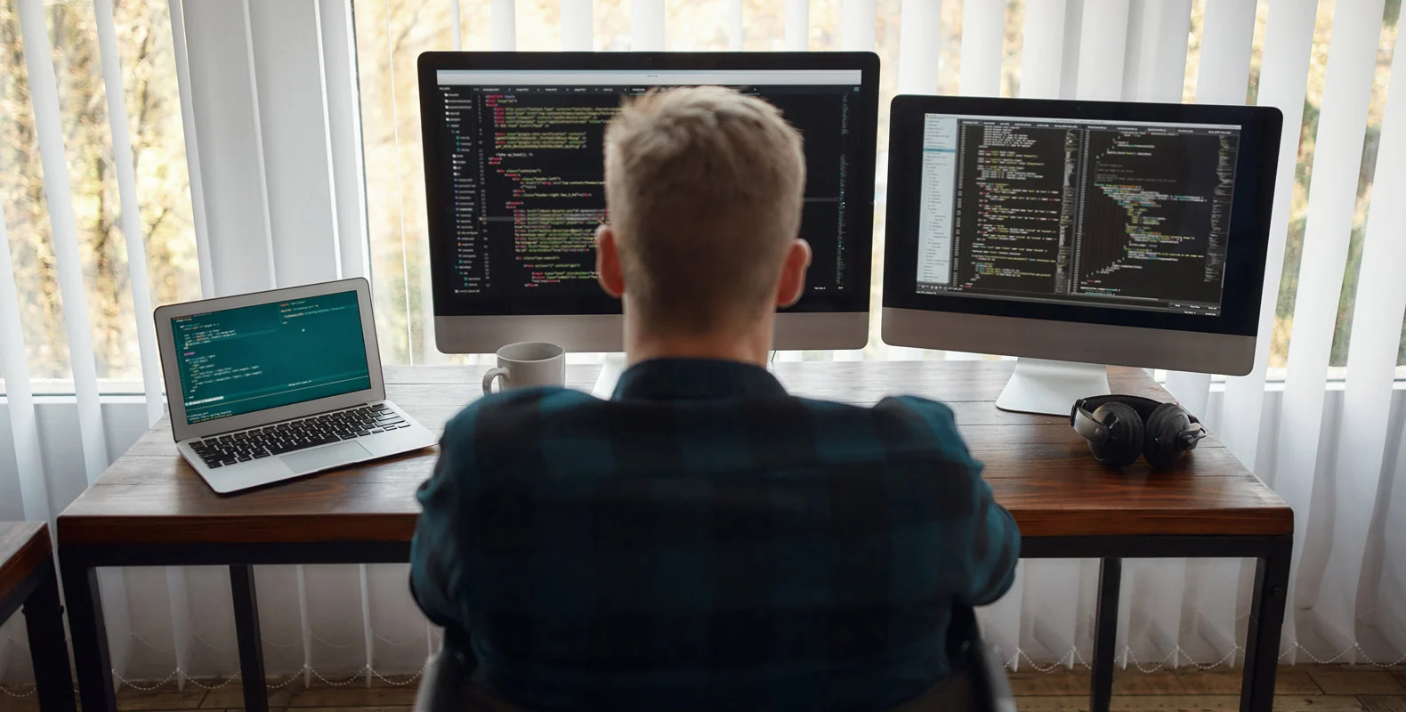 A person sits at a desk with a laptop and two monitors displaying code, in front of a large window with sheer curtains.