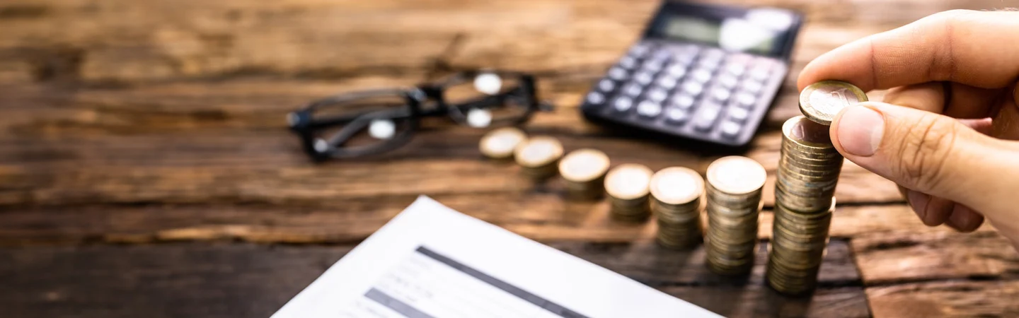 Close-up of hand stacking coins on wooden table, with calculator, glasses, and financial document in the background.