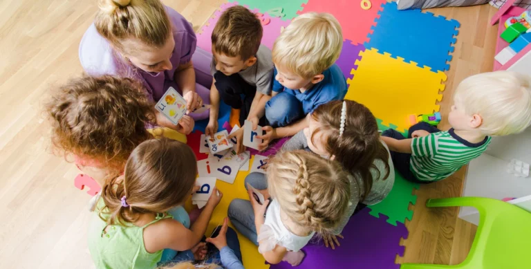 Children sitting in a circle on colorful mats, playing with alphabet flashcards, supervised by an adult.