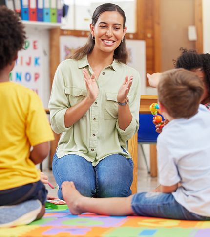 A smiling woman, likely a preschool teacher, sits on the floor clapping her hands, surrounded by young children in a colorful classroom—a lively scene of preschool education with shelves, books, and alphabet posters in the background.